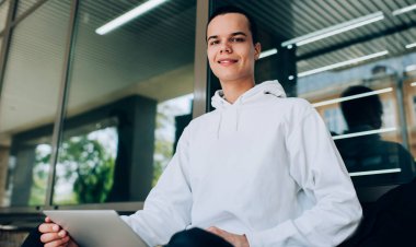 Mutlu, uzaktan kumandalı, dişlek tebessümle, modern binanın yanında tabletle oturup çalışırken kameraya bakarak mutlu bir erkek işçi.