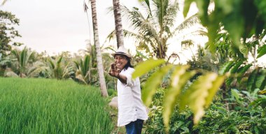 Şapkalı, neşeli erkek çiftçinin yarısı Ubud 'daki palmiye ağaçlarıyla pirinç tarlalarında el sallıyor ve el kol sallıyor. Neşeli Balili adam tarım ve seyahat etmeyi seviyor.