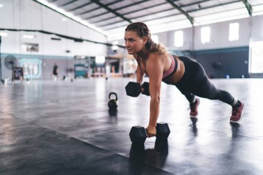 Spor giyimli güçlü bir beyaz atlet spor salonunda dambıl tutuyor, eşofmanlı bir kız yoğun çapraz formda vücut kaslarını çalıştırmak için sıkı ekipman kullanıyor.