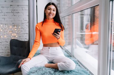 Gülümseyen Çinli kız pencere pervazında kürklü halıda oturuyor ve evde akıllı telefon kullanıyor. Ev eğlencesi ve eğlence. Genç çekici esmer kadın kameraya bakıyor. Geniş daire