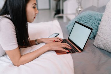 Akıllı telefonuyla yatakta yatan ve dizüstü bilgisayarında yazan Çinli bir kızın kırpılmış görüntüsü. Ev eğlencesi ve eğlence. Bin yıllık neslin güzel esmer kadını. Modern ferah daire
