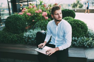 Siyah pantolonlu ve beyaz gömlekli düşünceli genç bir erkek dizüstü bilgisayarda klavyede otururken yüzünü çeviriyor.