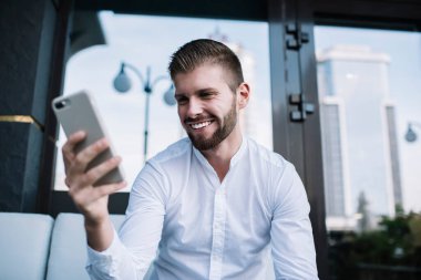 Beyaz tişörtlü, neşeli, sakallı genç bir erkeğin cep telefonuyla internette gezinip kafenin terasında oturmasından.