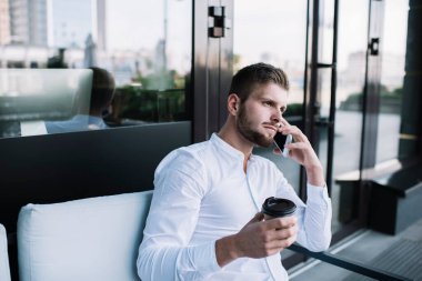 Düşünceli erkek girişimci, iş sırasında elinde bir fincan kahveyle bankta otururken ve cep telefonuyla konuşurken başka tarafa bakıyor.