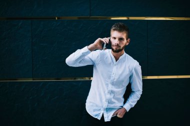 Kendine güvenen, beyaz tişört giyen, modern koridor duvarının yanında el ele duran, kendine güvenen, sakallı bir adam.