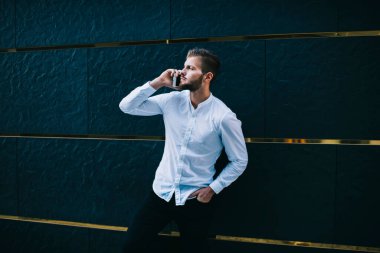 Ciddi bir yetişkin sakallı girişimci resmi giysiler içinde koyu mavi duvara yaslanmış, elini cebine atmış ve cep telefonuyla konuşuyor.