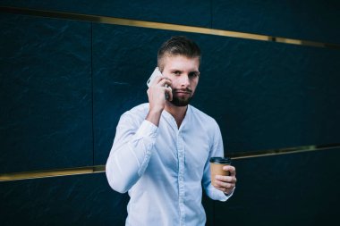 Ciddi sakallı, beyaz tişörtlü, akıllı telefonlu bir adam elinde bir bardak sıcak içecekle modern binanın duvarına bakarken kameraya bakıyor.