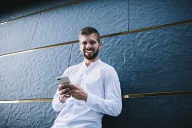 Resmî kıyafetli, kameraya bakıp gülümseyen, akıllı telefon kullanan ve modern binanın taş duvarına yaslanan pozitif sakallı adamın alçak açısı.