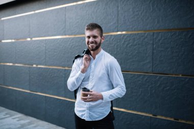 Genç, kendine güveni tam sakallı, ceketini omzunun üstünde tutan ve modern binanın yanında duran kağıt bardak sıcak içecekle kameraya bakan bir adam.