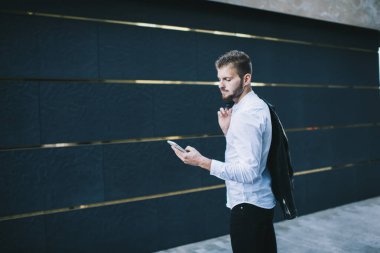 Moda kıyafetli, sakallı, cep telefonlu, modern bir binanın yanında duran yakışıklı bir adamın yan görüntüsü.