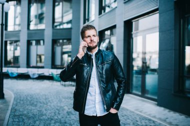 Moda kıyafetleri içindeki yakışıklı genç adam modern binanın yanındaki caddede konuşurken elini cebine atıyor.