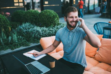 Gündüz vakti masada elinde kahve ve not defteriyle akıllı telefonla konuşurken terasta bilgisayarda çalışan serbest çalışan mutlu genç adam.