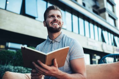 Düşük açılı mutlu sakallı, serbest çalışan genç bir erkek. Gözünü kaçırıyor ve kafeteryada oturup not tutuyor.