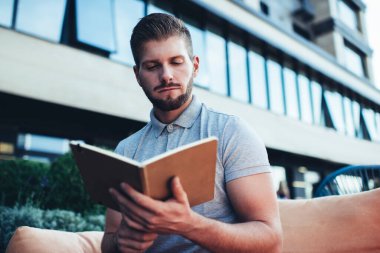 Ciddi sakallı, serbest çalışan, serbest giyinen, kafeteryada oturan ve ajandadaki randevularla ilgili notlar okuyan bir erkek.