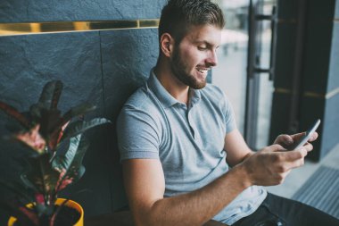 Neşeli, kendine güvenen, sıradan kıyafetli, modern binanın yanında cep telefonuyla mesajlaşan ve caddede saksıda yeşil bitki yetiştiren erkek.