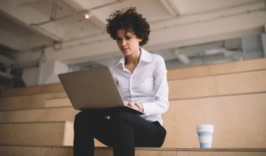 Milenyum Kafkas kızı dizüstü bilgisayarda yazıp izliyor. Modern başarılı kadın kavramı. Genç kıvırcık kadın ahşap bankta oturuyor. Akıllı telefon ve kahve fincanıyla birlikte çalışma odasında.