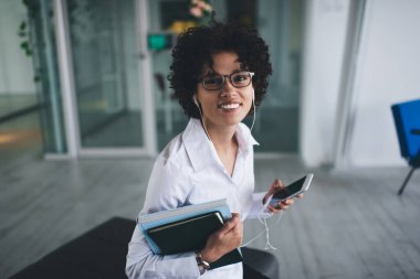 Milenyum Kafkasyalı iş kadını kulaklıkla kulaklıkla müzik dinliyor ve kameraya bakıyor. Modern başarılı kadın kavramı. Ofiste açık alanda gözlüklü, gülümseyen kıvırcık kız.