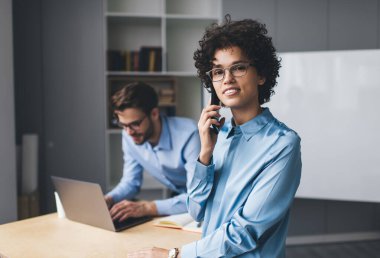 İş kadını akıllı telefondan konuşuyor ve erkek arkadaşı masada dizüstü bilgisayarda yazarken kameraya bakıyor. Modern başarılı insanlar kavramı. Genç Kafkas milenyum insanları ofiste.