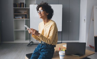 Milenyum Kafkasyalı iş kadını cep telefonu tutuyor ve iş sırasında başka tarafa bakıyor. Modern başarılı kadın kavramı. Açık uzay ofisindeki tahta masada gözlüklü, gülümseyen kıvırcık kız.