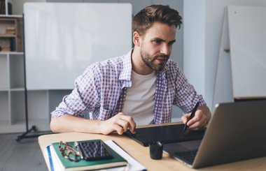 Milenyum Avrupalı işadamı dizüstü bilgisayardan izliyor ve grafik tabletine bir şeyler yazıyor. Modern başarılı adam kavramı. Açık uzay ofisindeki masada oturan genç odaklı sakallı adam.