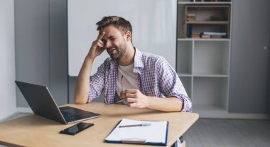 Bin yıllık Avrupalı iş adamının dizüstü bilgisayarla çalışırken başı ağrıyor. Modern başarılı adam kavramı. Açık uzay ofisindeki ahşap masada oturan gözlüklü genç adam.