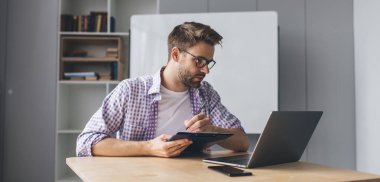 Milenyum Kafkas işadamı dizüstü bilgisayardan izliyor ve panoya bir şeyler yazıyor. Modern başarılı adam kavramı. Gözlüklü genç adam açık uzay ofisinde masada oturuyor.