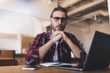 Optik gözlüklü ciddi bir erkek portresi. Uzaktan kumandalı iş sırasında kameraya bakıyor. Kafkas bilişim uzmanı dizüstü bilgisayarla poz veriyor.