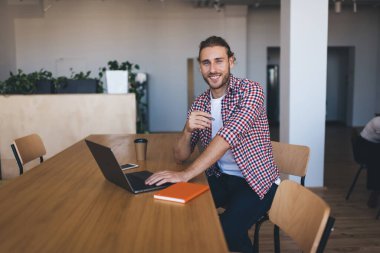 Milenyum Kafkas işadamı laptopta çalışırken kameraya bakıyor. Modern başarılı adam kavramı. Genç gülümseyen erkek girişimci açık uzay ofisindeki ahşap masada oturuyor.