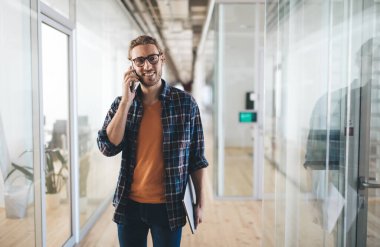 Milenyum Kafkasyalı iş adamı laptopuyla iş sırasında akıllı telefondan konuşuyor. Modern başarılı adam kavramı. Genç sakallı, gülümseyen erkek girişimci gözlüklü, açık uzay ofisinde.