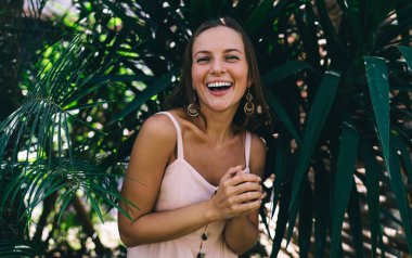 Mutluluktan kahkahalarla gülen ve neşeyle sevinen neşeli kadın turistin yarım boy portresi. Tropikal bir ortamda bitkilerle poz verirken. Kameraya gülümseyen samimi bir model.