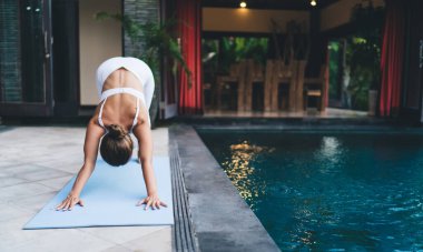 Calm hipster girl stretching back for training body muscles during patio workout on blue mat, active female athlete in sportswear doing exercise practicing for stretching flexibility at pool terrace clipart