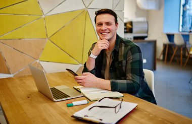 Genç gülümseyen adam dizüstü bilgisayarla masada otururken cep telefonunu elinde tutuyor. Çevrimiçi olarak çalışmak, teknolojiyi kullanmak 