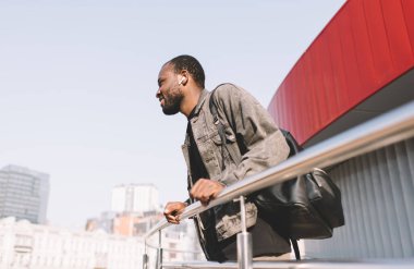 Seyahat sırt çantalı Afro-Amerikalı turist elektronik kulaklıklardan kaydedilen sesleri dinliyor, milenyum koyu tenli kulaklıklı hippi adam şehir gezileri için zaman harcıyor.