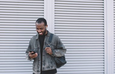 Bluetooth kulaklıklı neşeli koyu tenli adam sosyal medyada ağ takipçileriyle sohbet ederken pozitif müzik dinliyor, Afrikalı Amerikalı seyahat blogcusu cep telefonundan çalma listesinin keyfini çıkarıyor