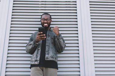 Neşeli Afrikalı Amerikalı seyahat blogcusunun yarım boy portresi. Gündüz vakti online zaferden memnun kalan, görme düzeltmesi için optik gözlüklü neşeli hipster adam elinde cep telefonu tutuyor.