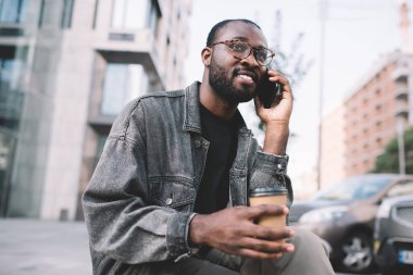 Klasik göz giysisi içinde koyu tenli bir adam akıllı telefon uygulaması ile arıyor. Kentsel şehirde 4G kullanıyor. Optik gözlüklü Afro-Amerikan hippi adam cep telefonu teknolojisiyle arama yapıyor.