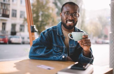 Beyaz fincan sıcak kahveyle gezen Afro-Amerikan hipster adamın portresi. Şehirde dinlenmek için kameraya gülümsüyor. Optik gözlüklü neşeli bir erkek, göz düzeltmesi için poz veriyor.