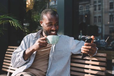 Elektronik kulaklıklı mutlu koyu tenli adam cep telefonundan Bluetooth bağlantısı kullanarak cep telefonundan gelen e-postayı kontrol ediyor kulaklıklı neşeli hippi adam video izliyor.