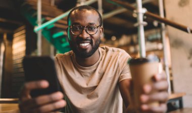 Hücresel cihaz kullanan ve boş zamanlarında kameraya gülümseyen neşeli hippi portresi. Optik gözlüklü mutlu Afro-Amerikan adam. Bulanık akıllı telefon cihazı ve kahve tutuyor.