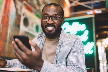 Klasik gözlüklü neşeli erkek öğrencinin portresi kameraya gülümserken kapalı alanda akıllı telefon uygulaması kullanıyor, modern mobil cihazlı mutlu Afro-Amerikan hipster adam cep telefonunun keyfini çıkarıyor.