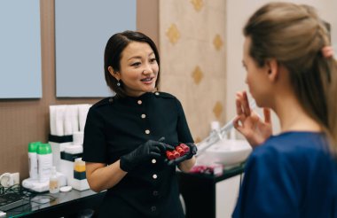 Güzellik uzmanı, güzellik salonundaki beyaz kadın müşteriyi seçmek ve dinlemek için kavanozları rujla gösteriyor. Kalıcı makyaj dudakları prosedürü. Yetişkin Asyalı kadın üstat eldiven giyiyor. Güzellik konsepti