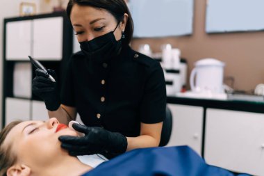 Güzellik uzmanı, kuaförde genç beyaz kadın müşterinin iğnesiyle kalıcı dudak ameliyatı yapıyor. Yetişkin Asyalı kadın üstat eldiven ve tıbbi eldiven giyiyor. Güzellik konsepti