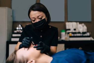Güzellik uzmanı kuaförde gözü kapalı beyaz bir kadının iğnesiyle kalıcı makyaj dudak ameliyatı yapıyor. Eldiven ve tıbbi maske takan Asyalı kadın üstat. Güzellik konsepti