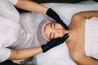 Kız yüzüne masaj yapan ve modern güzellik salonundaki kameraya bakan kadın güzellik uzmanının en iyi görüntüsü. Kapalı gözlü, tıbbi şapkalı genç Asyalı kadın müşteri. Vücut ve cilt bakımı kavramı