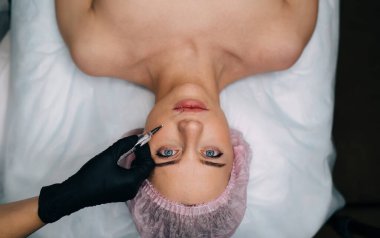 Güzellik uzmanının güzellik salonundaki kıza şırıngayla botoks yaparken kısmi görüntüsü. Avrupalı genç bayan müşterinin kameraya bakan tıbbi şapkalı hâli. Güzellik konsepti. Yüz gerdirme