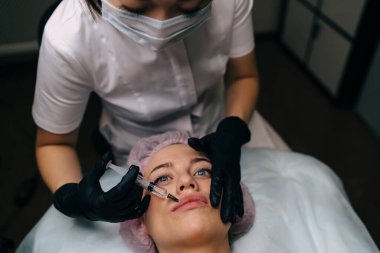Güzellik uzmanının güzellik salonundaki kıza şırıngayla botoks yaparken çekilmiş görüntüsü. Tıbbi başlık takan beyaz bir kadın müşteri. Güzellik konsepti. Eldiven ve tıbbi maske takan kadın bir usta.