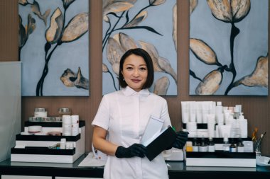 Kadın güzellik uzmanının elinde kozmetik ürünlerle kameraya baktığı ön manzara. Eldiven takan gülümseyen yetişkin Asyalı kadın kozmetik uzmanı. Güzellik konsepti. Modern güzellik salonunun içi.