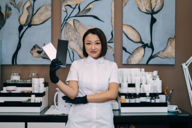 Kadın güzellik uzmanının kozmetik ürünleriyle paketleri göstermesi ve kameraya bakması. Eldiven takan gülümseyen yetişkin Asyalı kadın kozmetik uzmanı. Güzellik konsepti. Modern güzellik salonunun içi.