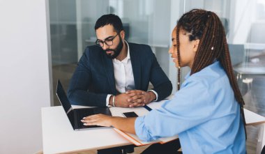 Orta Doğulu erkek patron, genç bayan çalışanla ofis toplantısı sırasında modern dizüstü bilgisayarda vizyon düzeltmesi yapmak için video sunumu yapmak ve beyin fırtınası yapmak için optik gözlük takıyor.