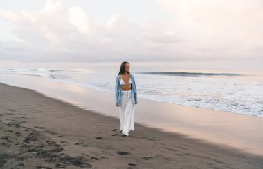 Muhteşem beyaz kadın plajda sakin ve kaygısız bir şekilde yürürken tatilin tadını çıkarıyor. Beyaz yaz giysileri içinde güzel, sağlıklı bir kadın tatil boyunca deniz kıyısında gezer.